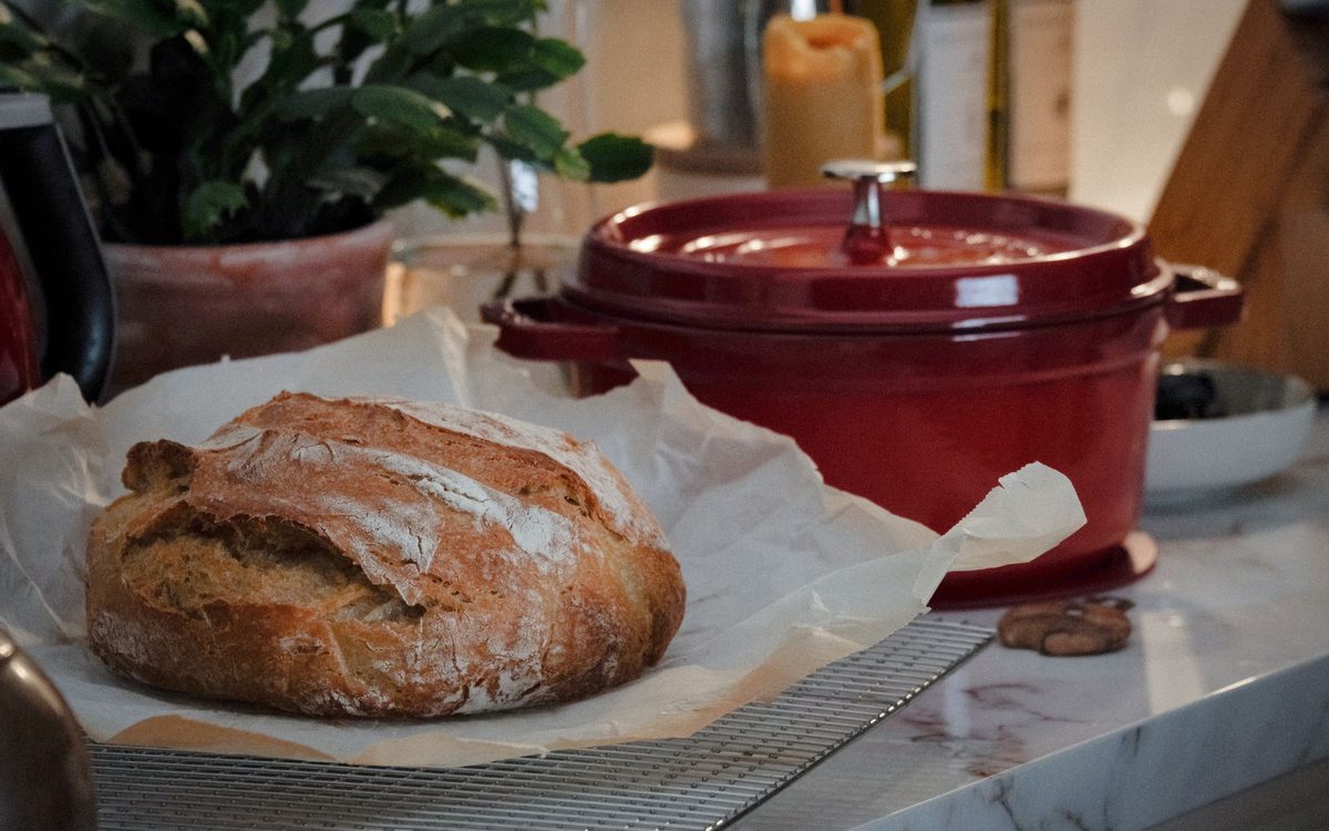 Close-up of a rustic loaf of bread on a kitchen counter with a red Dutch oven nearby.